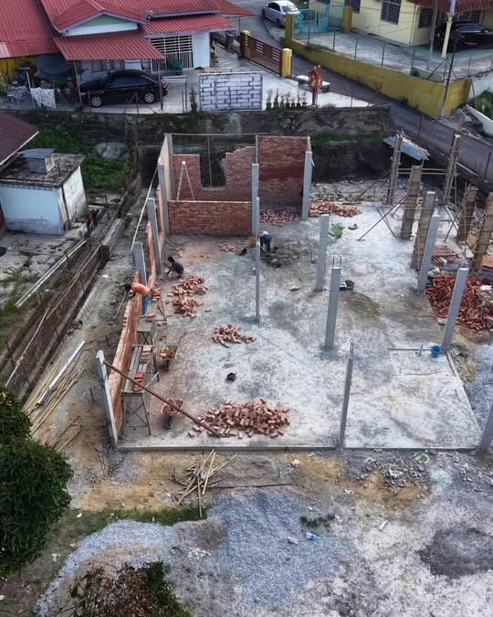 A high-angle drone view of an early-stage residential build: a poured ground-floor slab, freshly cast concrete columns, part-built red brick walls, stacked brick pallets and timber formwork.