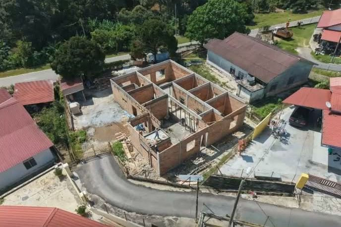 A high-angle drone view of a single-storey house shell at brickwork stage beside a road, with internal room divisions legible as brick walls and materials stacked around the perimeter.
